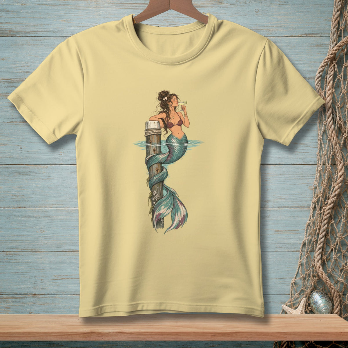 Light yellow t-shirt with a mermaid illustration holding a drink, leaning on a wooden post.