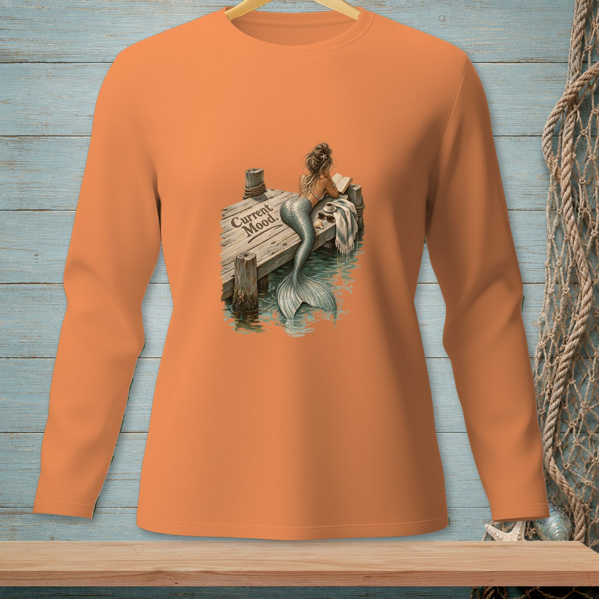 Orange long-sleeve shirt with a mermaid reading on a dock, saying "Current Mood."