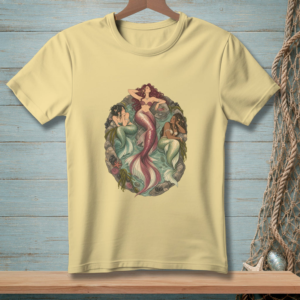 Three mermaids are depicted in a circular frame on a light yellow t-shirt.