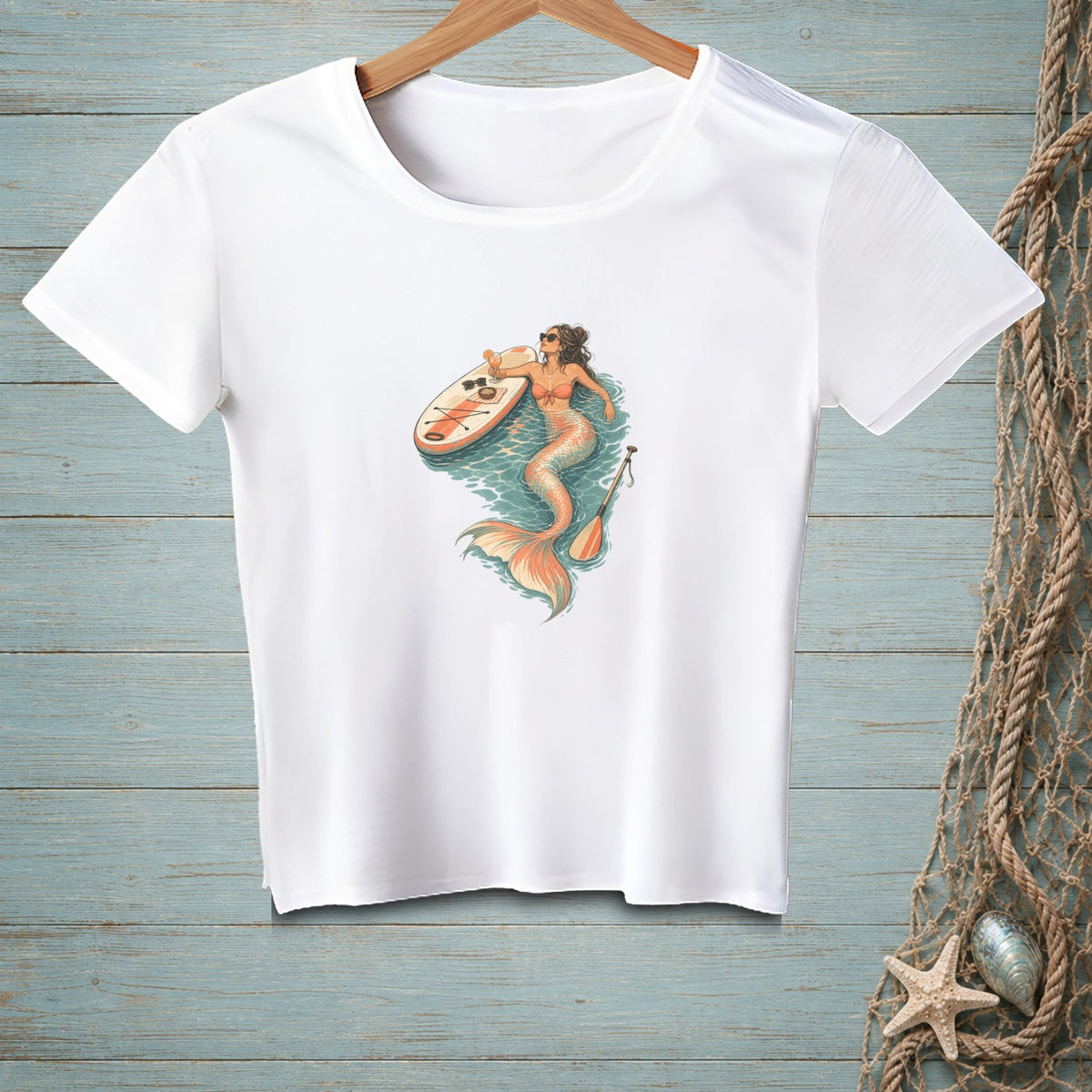 White t-shirt with a graphic of a mermaid on a paddleboard with sunglasses and a paddle.