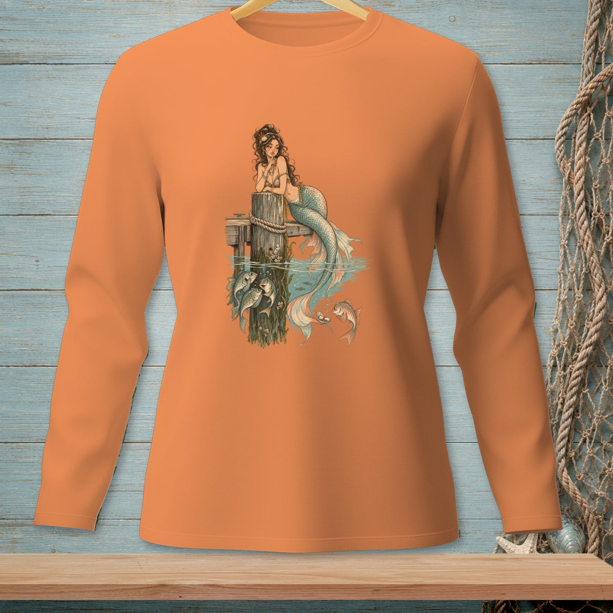 Orange long-sleeve shirt featuring a mermaid and fish graphic on a pier.