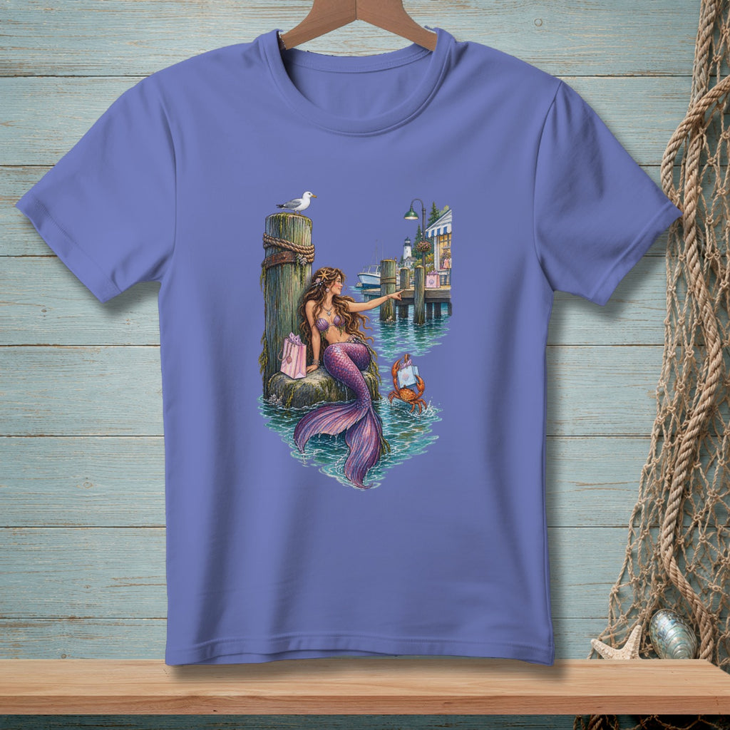 Mermaid sitting on pier with gift, crab, and lighthouse in background.