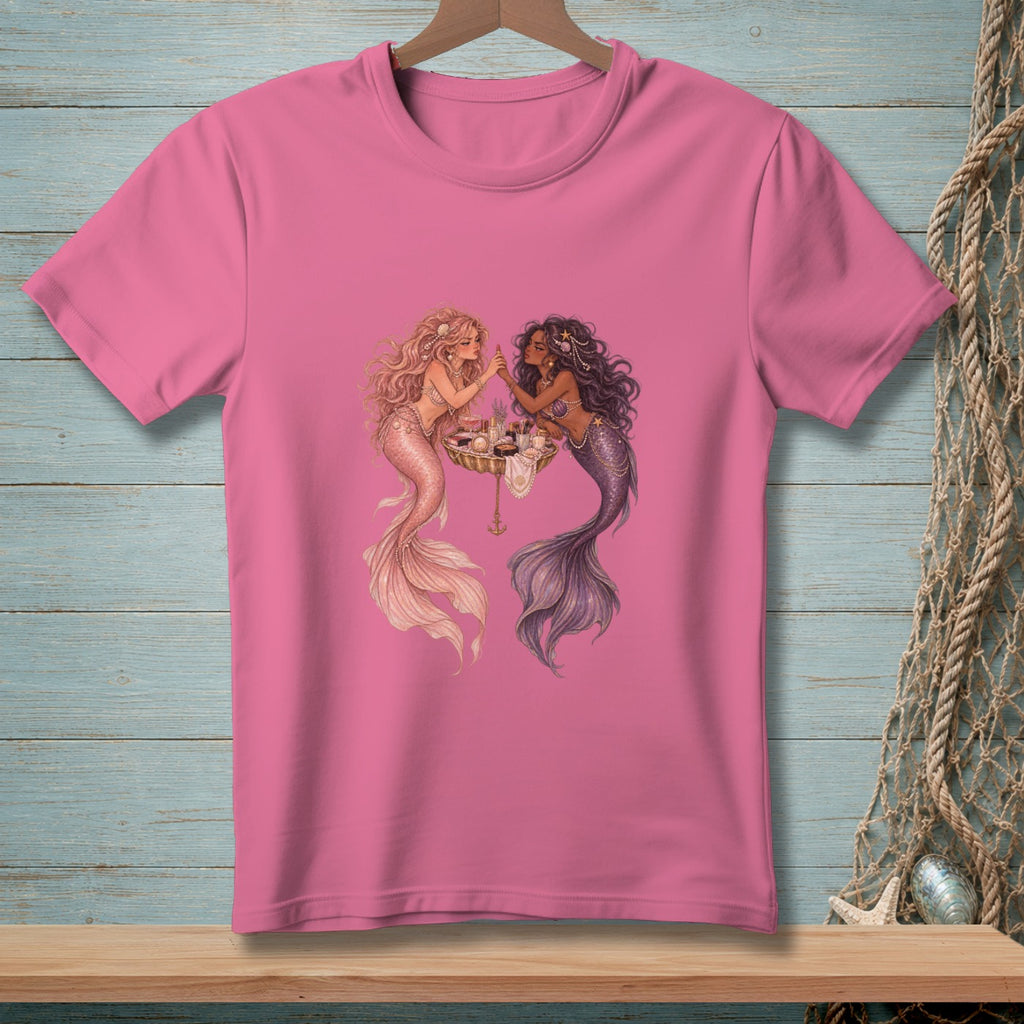 Pink t-shirt with two mermaids applying makeup around a table.