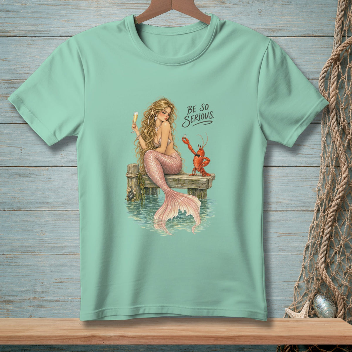 Mermaid with lobster on pier, text "BE SO SERIOUS" on light blue t-shirt.