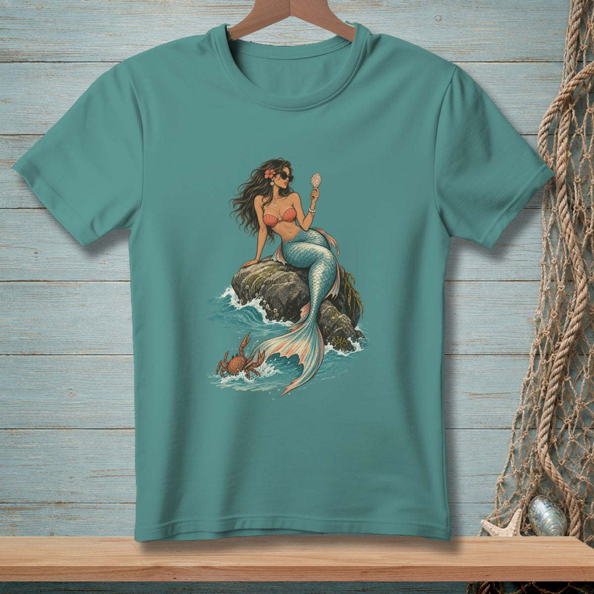 Teal t-shirt with a mermaid sitting on a rock, holding a shell, with a crab in the water.