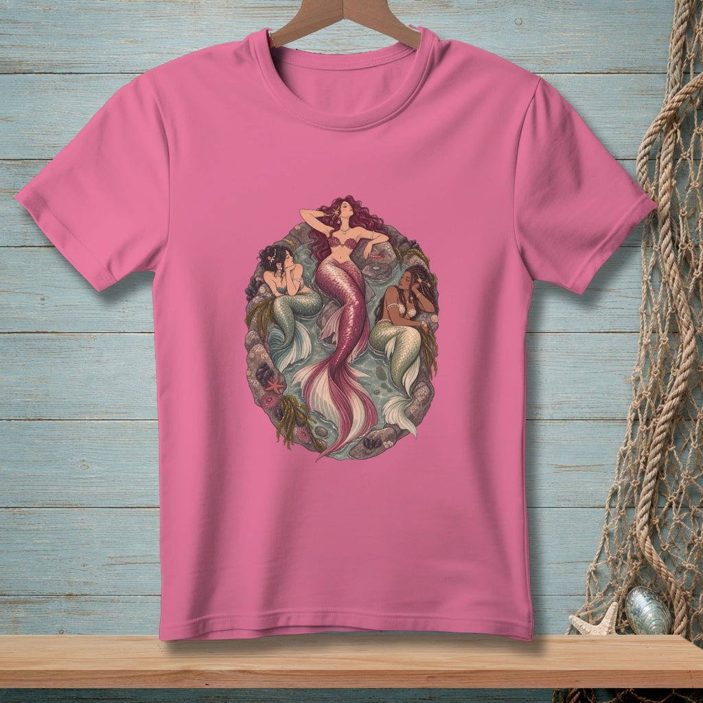 Pink t-shirt with three mermaids lounging on rocks in an oval shape.