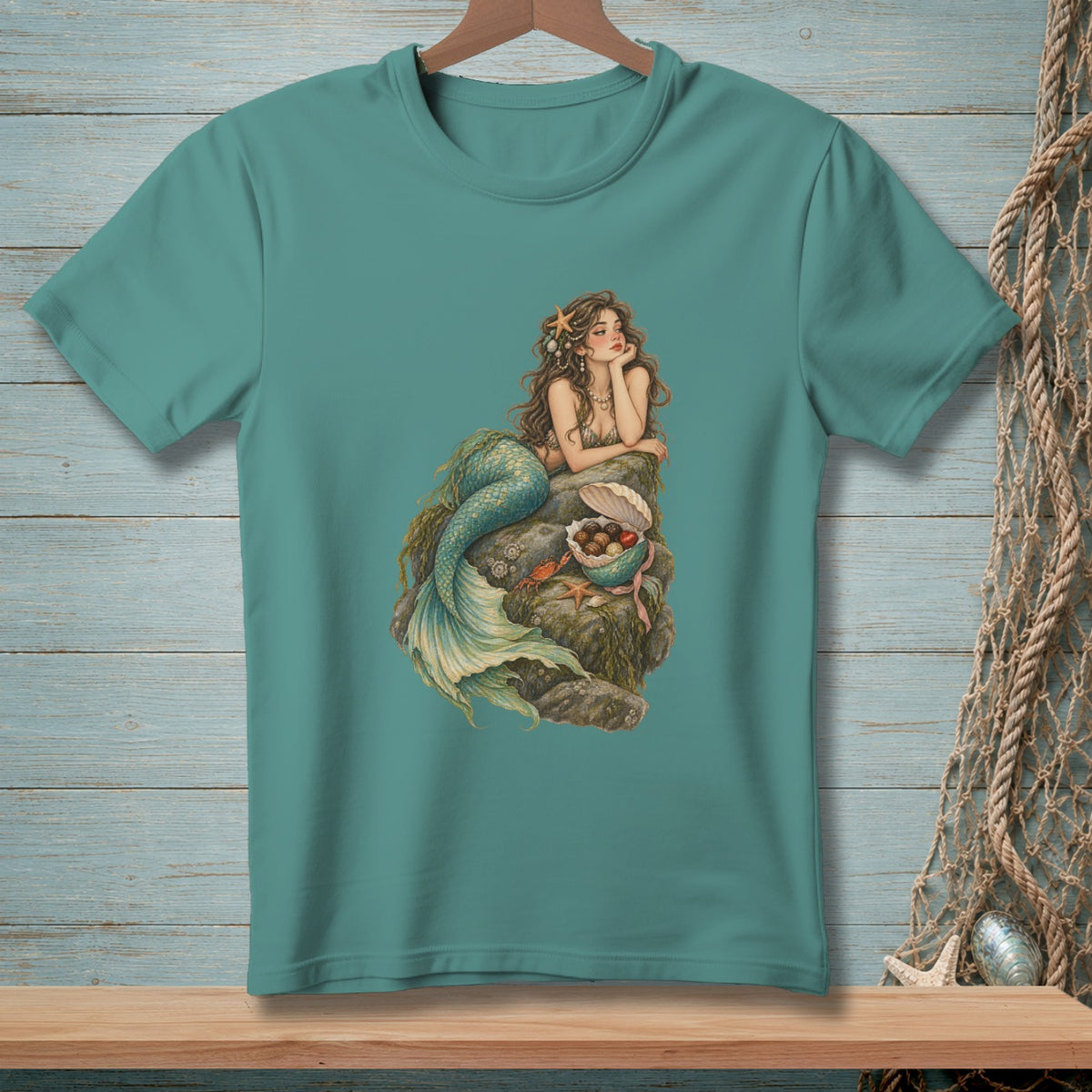 Teal t-shirt with a mermaid resting on rocks surrounded by shells and ocean treasures.