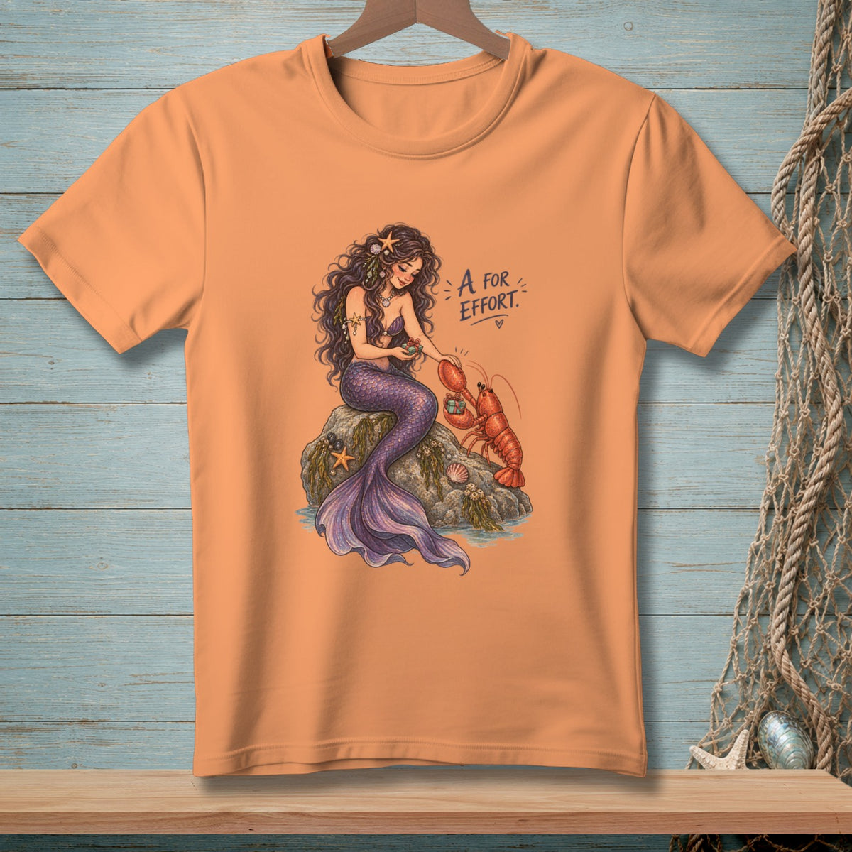 Peach t-shirt with illustration of a mermaid and lobster with text "A for Effort."