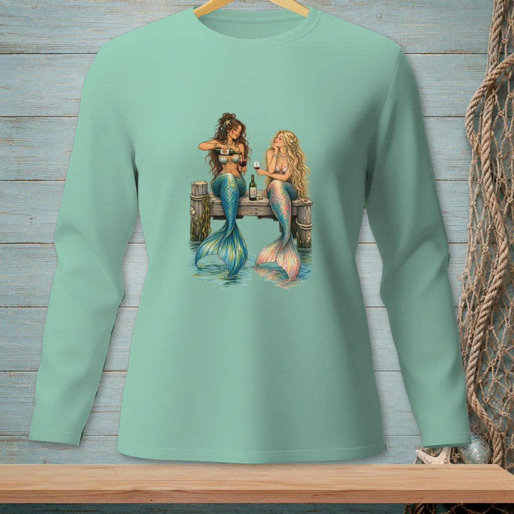 Two mermaids on a dock share wine with a bottle and glasses.