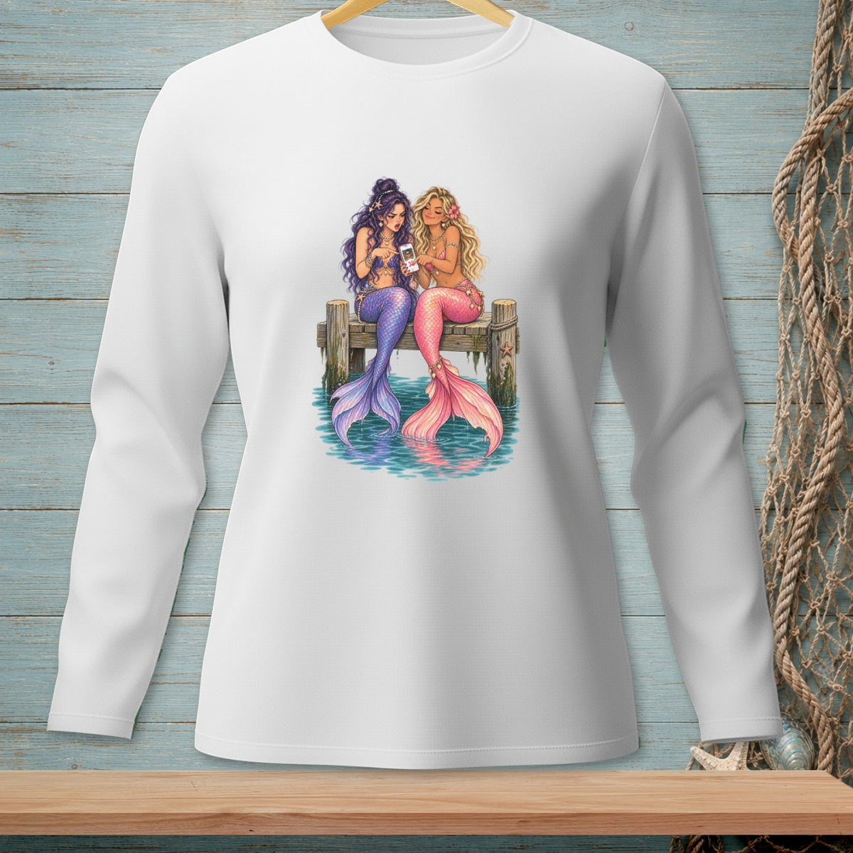 Two mermaids with purple and pink tails sit on a dock, looking at a smartphone.