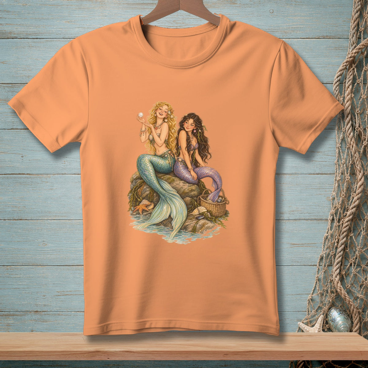 Orange t-shirt featuring two mermaids with tails of teal and purple, sitting on rocks with a pearl and basket.