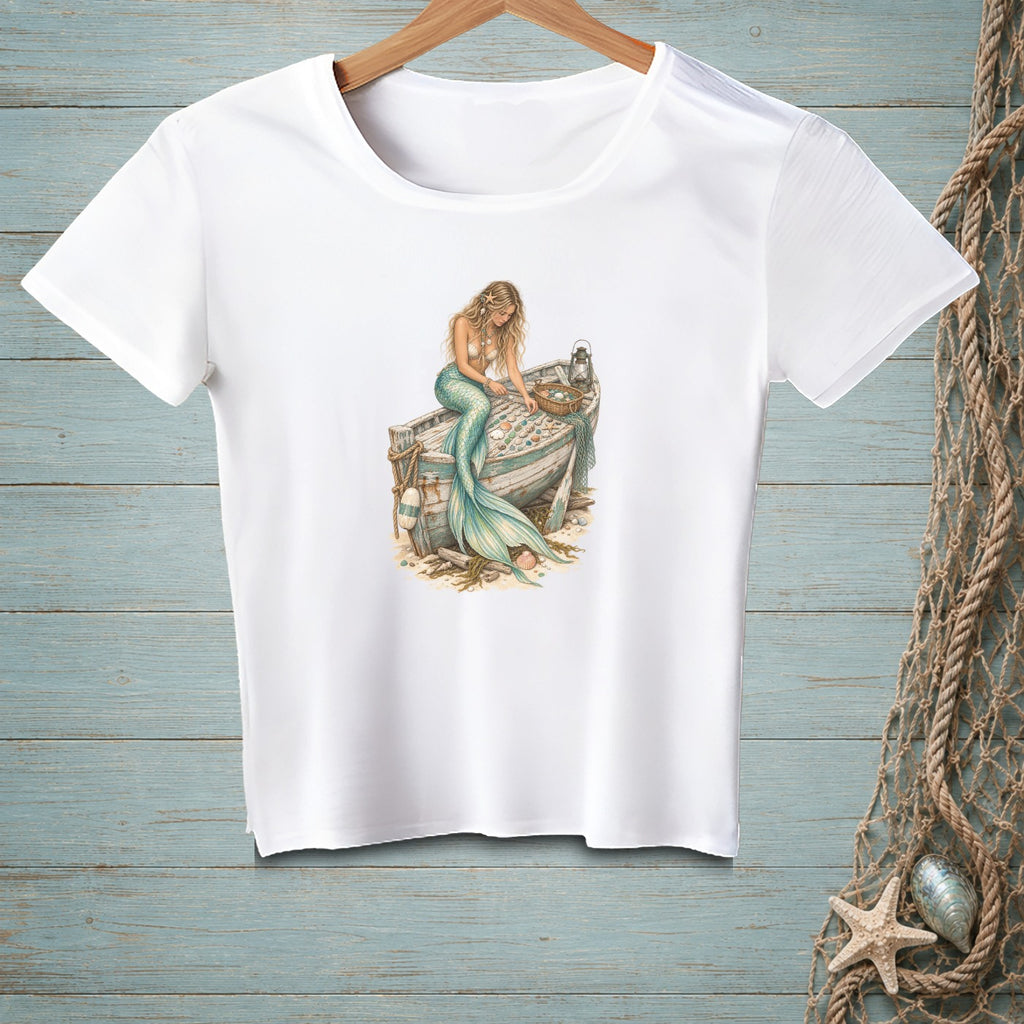 White t-shirt featuring a blonde mermaid sitting on a weathered boat filled with shells.