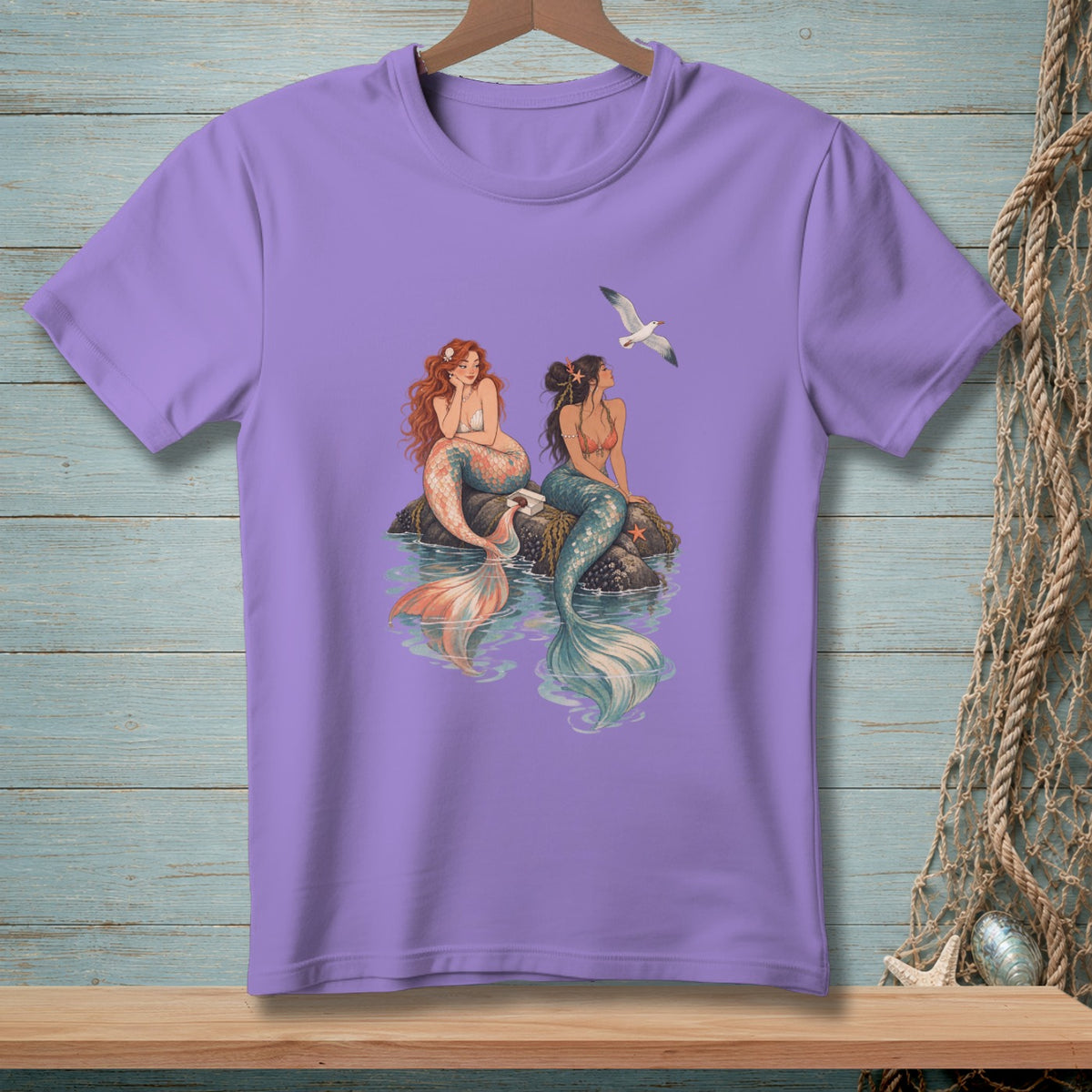 Lavender t-shirt with a graphic of two mermaids sitting on rocks with a seagull flying overhead.