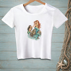 White t-shirt with two mermaids by a rock and blue water.