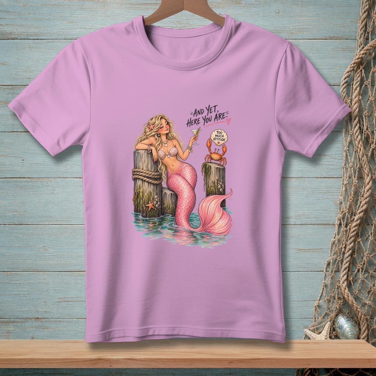 Pink t-shirt with a mermaid holding a drink, text "And yet, here you are", and a crab.