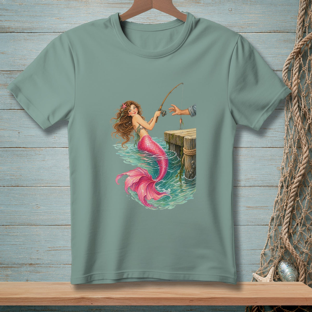 Green t-shirt with mermaid fishing from a dock with a bright pink tail.
