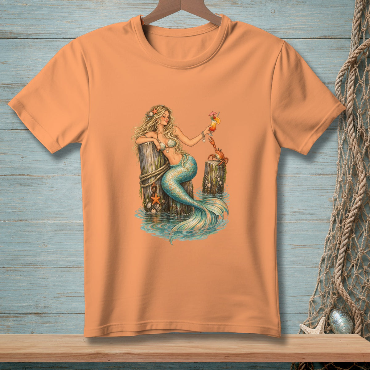 Peach t-shirt featuring a mermaid with a cocktail and a crab by a wooden post.