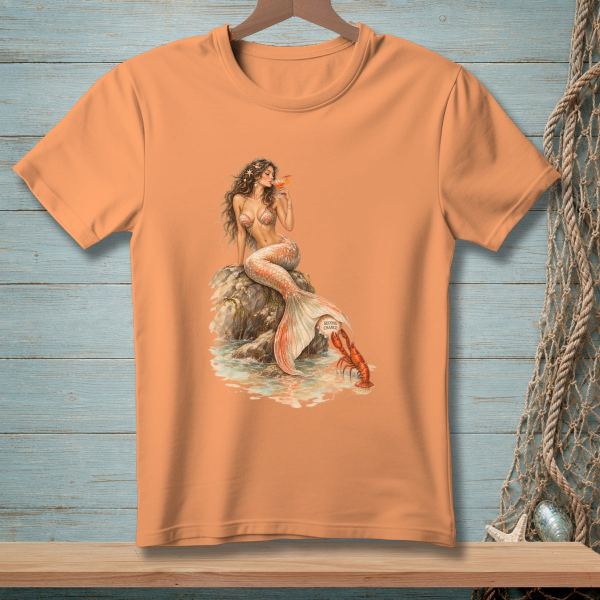 Peach t-shirt with watercolor illustration of mermaid drinking, sitting on a rock near a lobster.