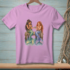 Two mermaids with colorful tails sit on a wooden dock, sharing a drink.