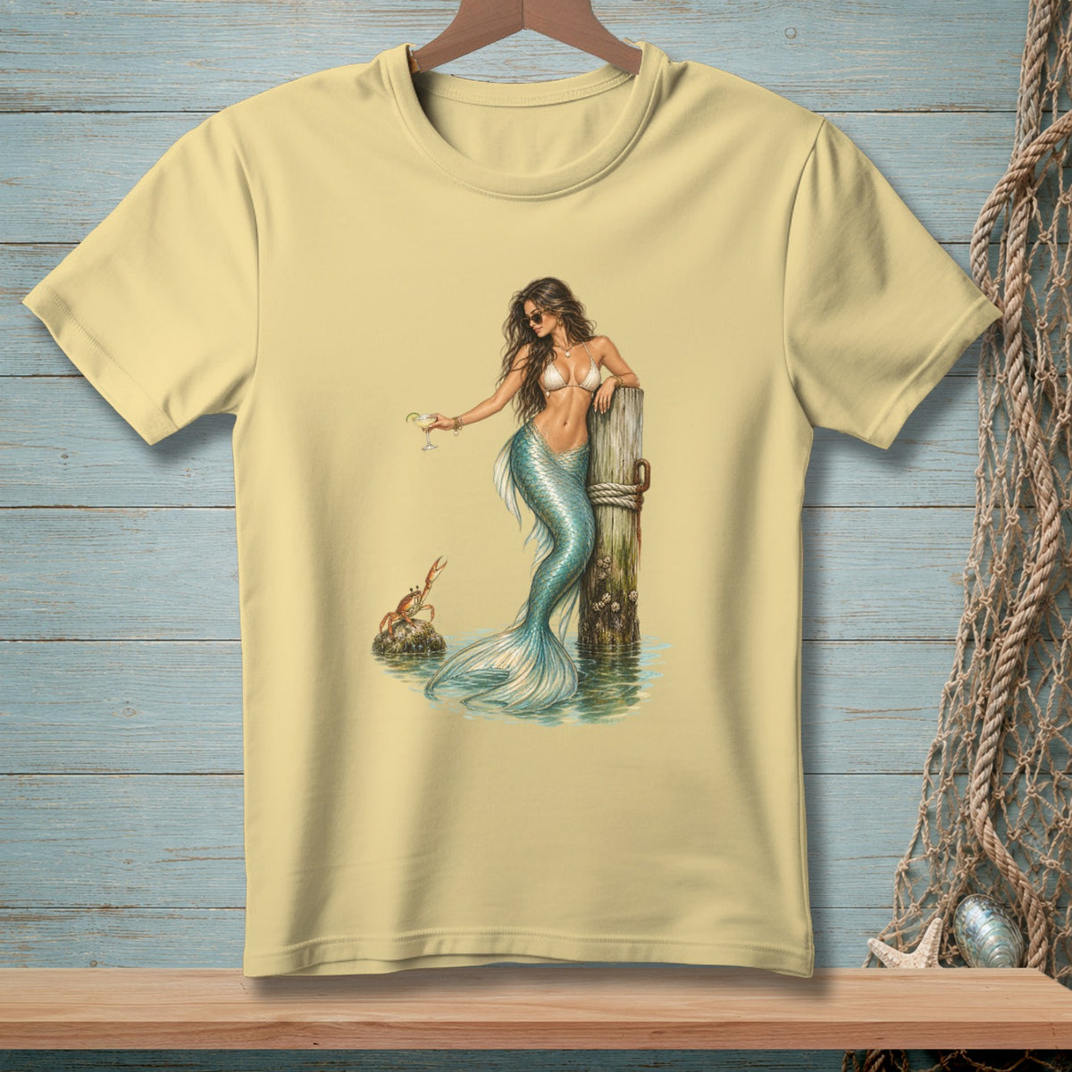 Pale yellow t-shirt with a mermaid holding a martini glass and a crab on a rock.