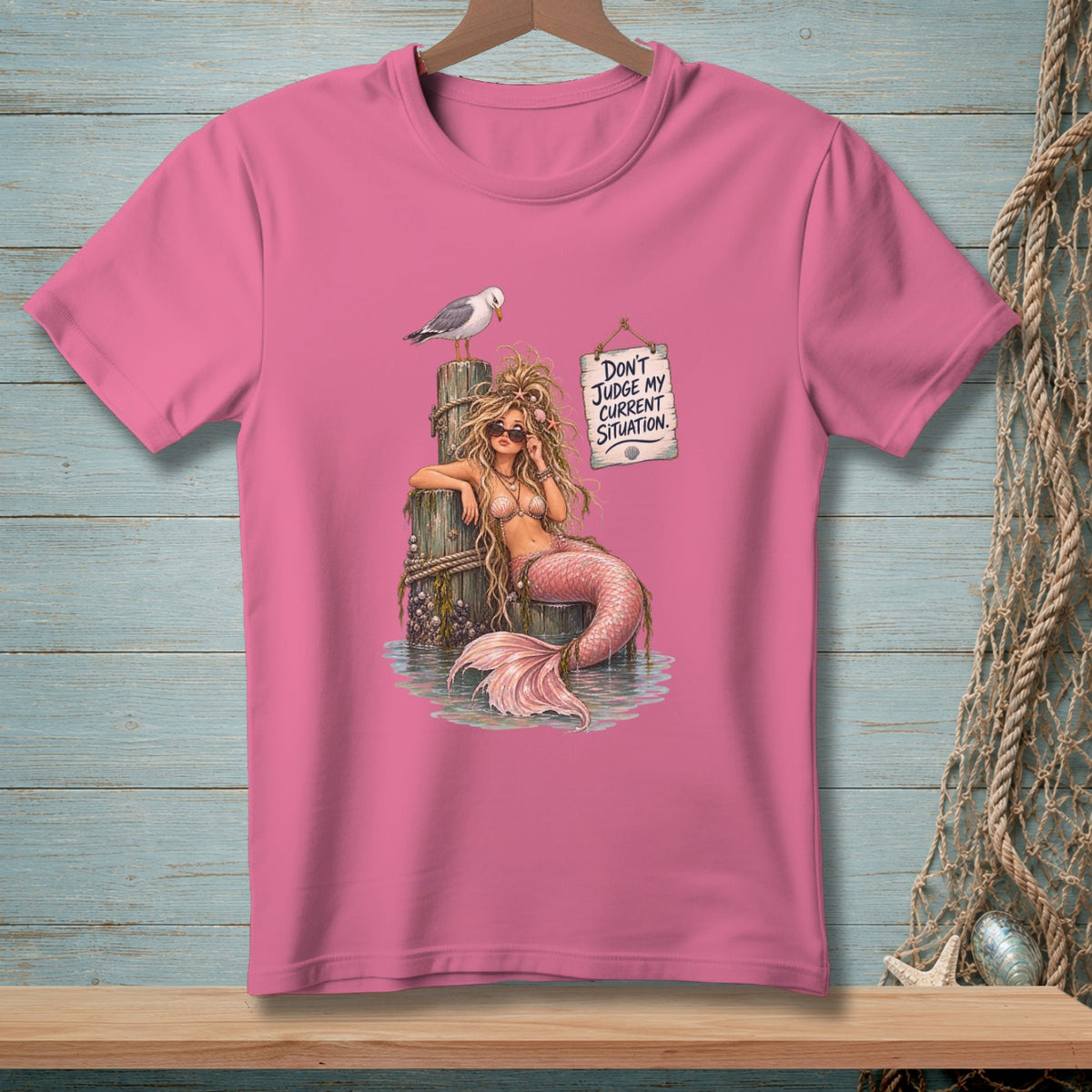 Pink t-shirt with a mermaid sitting on a pier with a seagull and a sign.