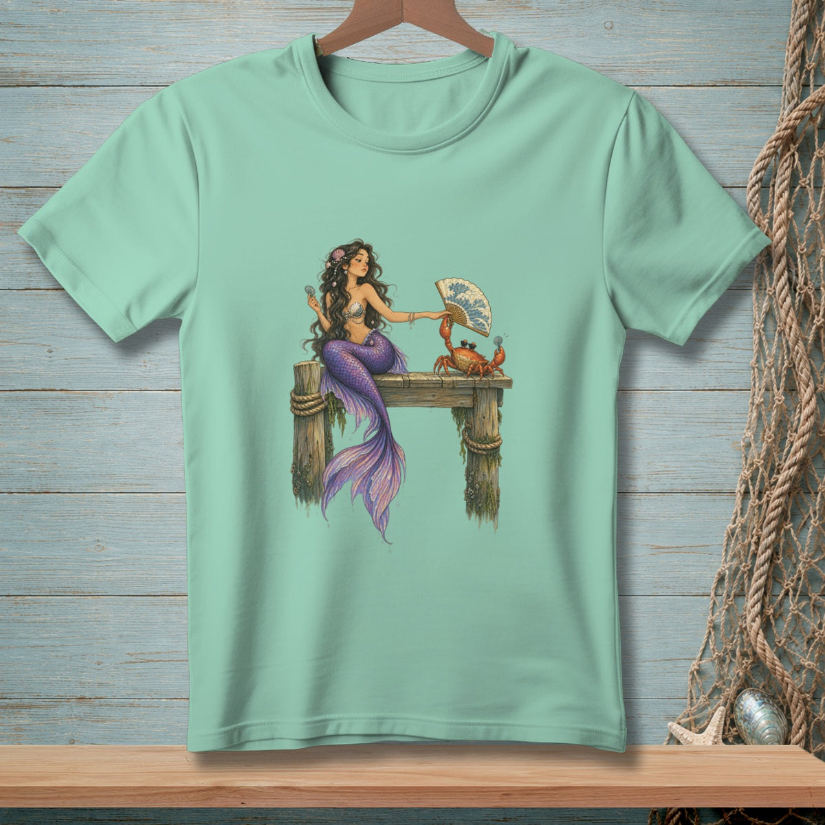 Mermaid and crab on a dock holding a fan, illustration on a light blue t-shirt.
