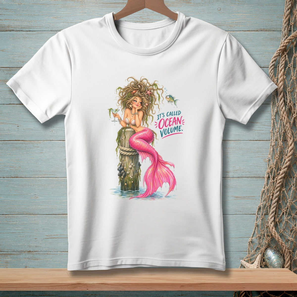 White t-shirt with a mermaid sitting on a wooden post, text reads "It's called Ocean Volume."
