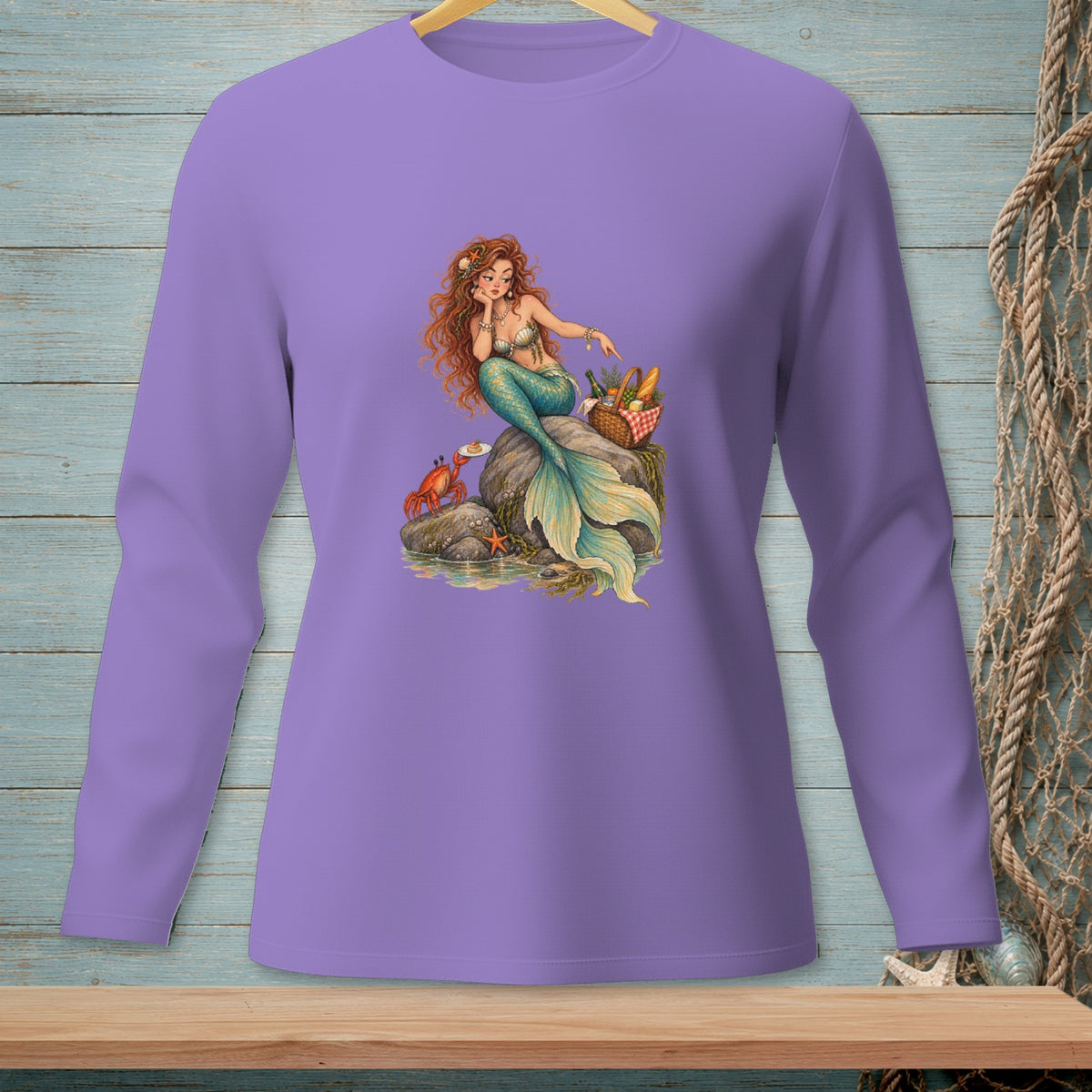 Illustration of a mermaid on a rock with a crab and picnic basket on a purple long-sleeved shirt.