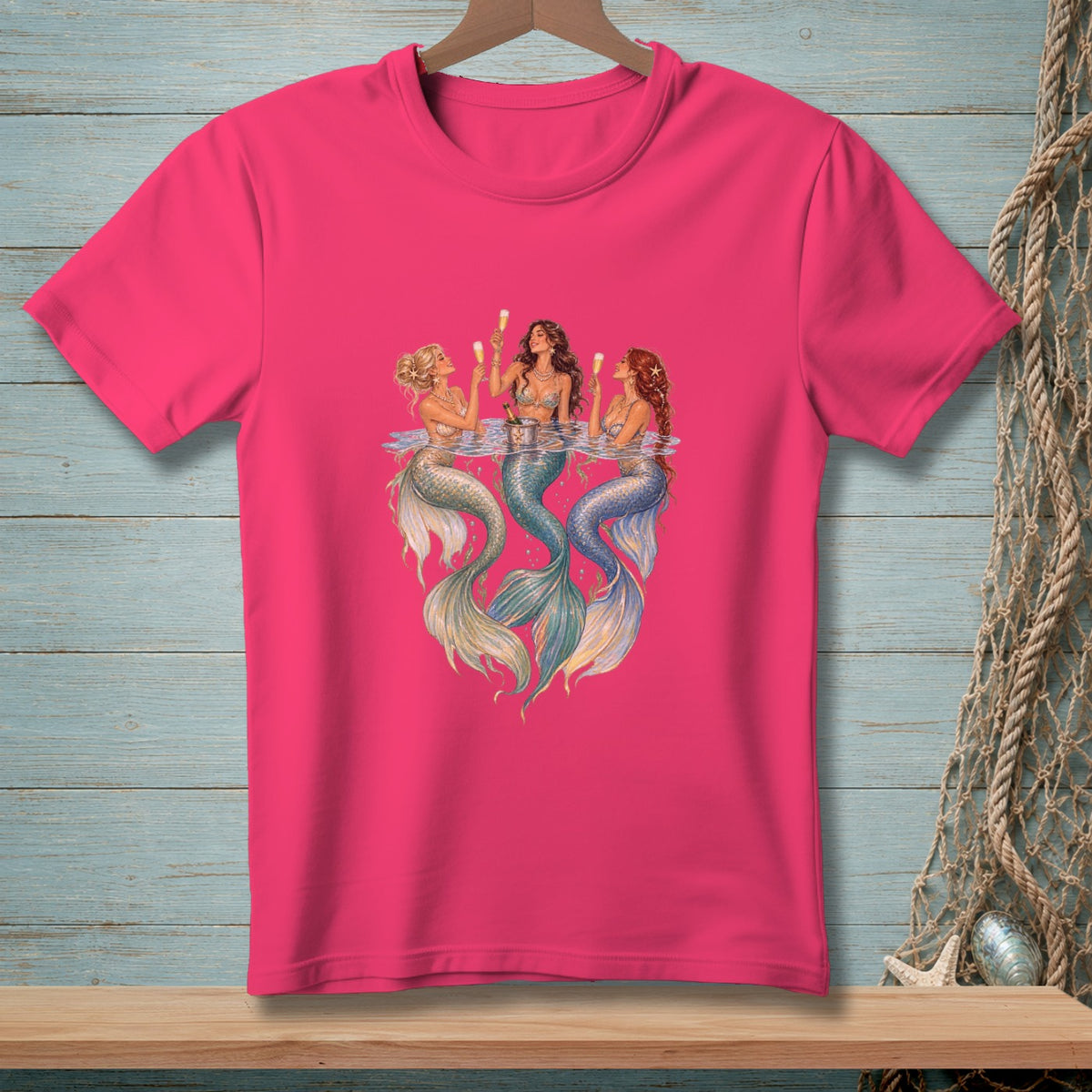 Three mermaids with champagne glasses sit in water on a pink t-shirt.