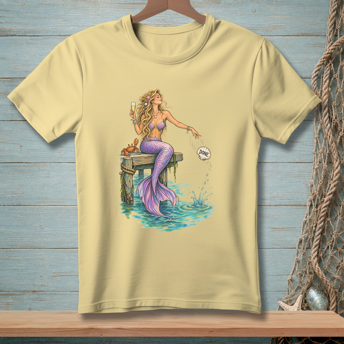 Light yellow t-shirt with a mermaid holding a champagne glass and a crab on a dock.