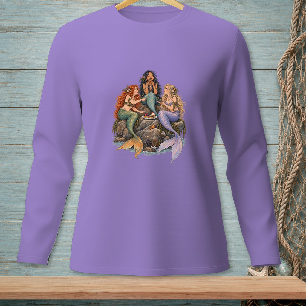 Three mermaids sitting on rocks sharing a cake and treats by the water.