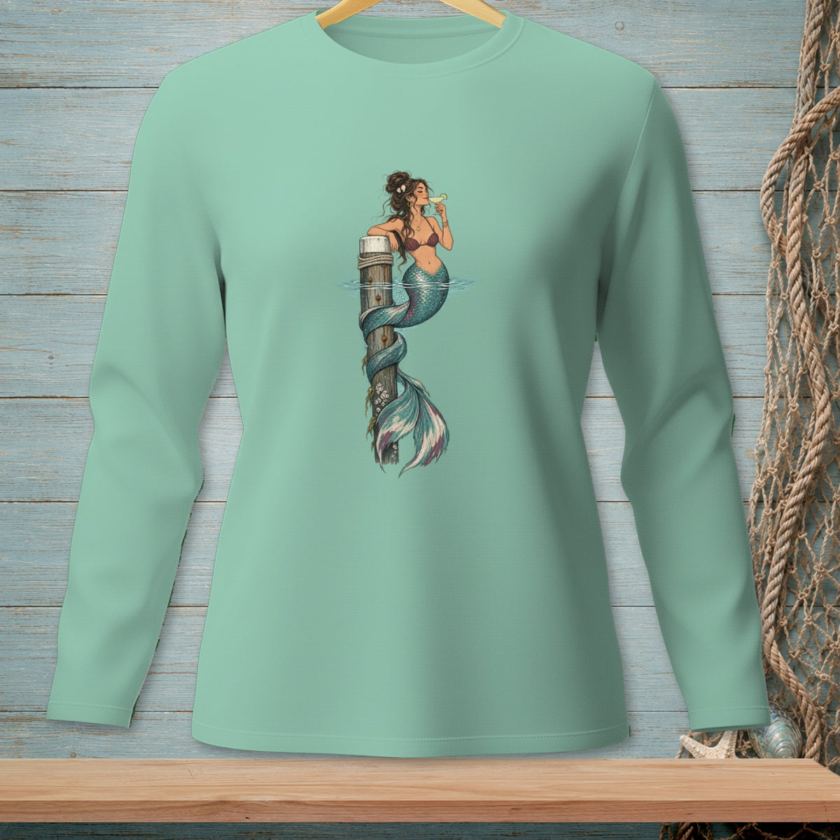 Mint long sleeve shirt with mermaid illustration drinking a cocktail on a post.