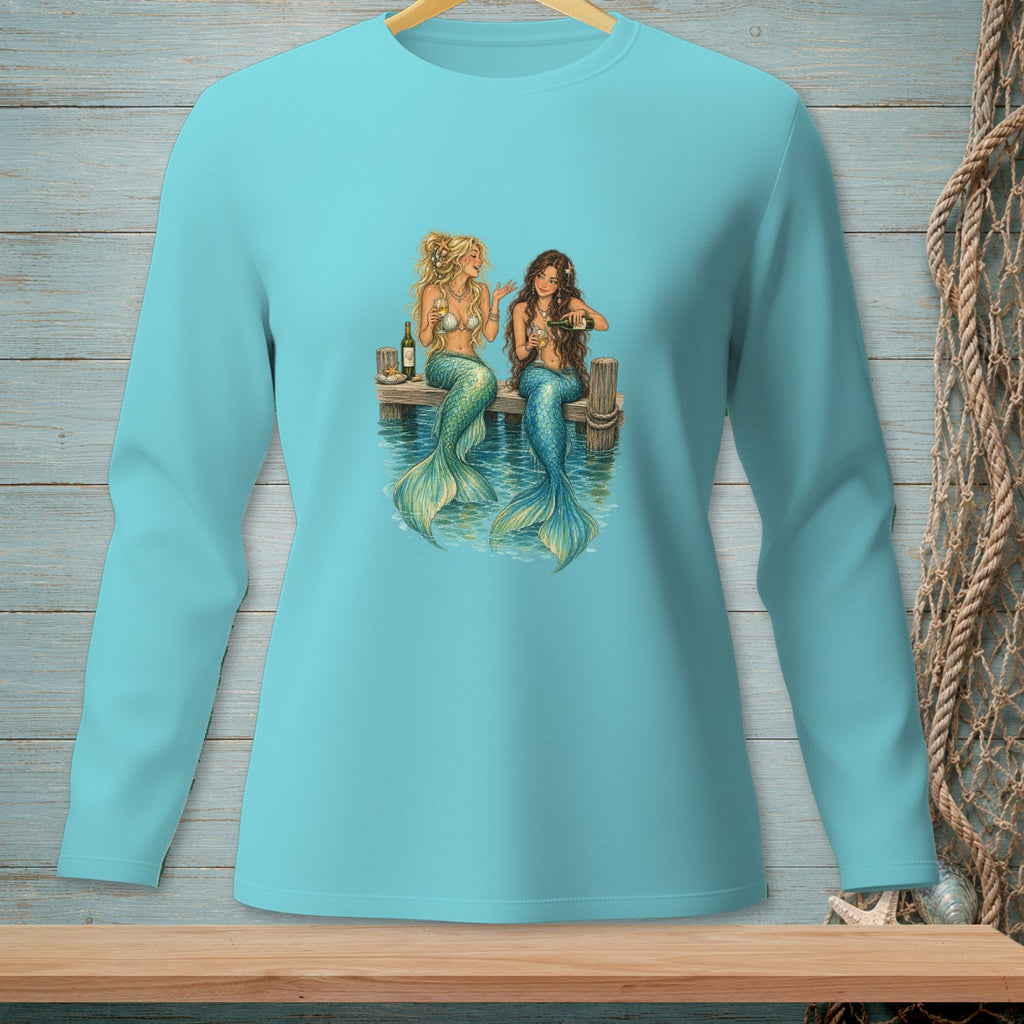 Two mermaids with shimmering tails sit on a wooden dock, holding glasses and a bottle.