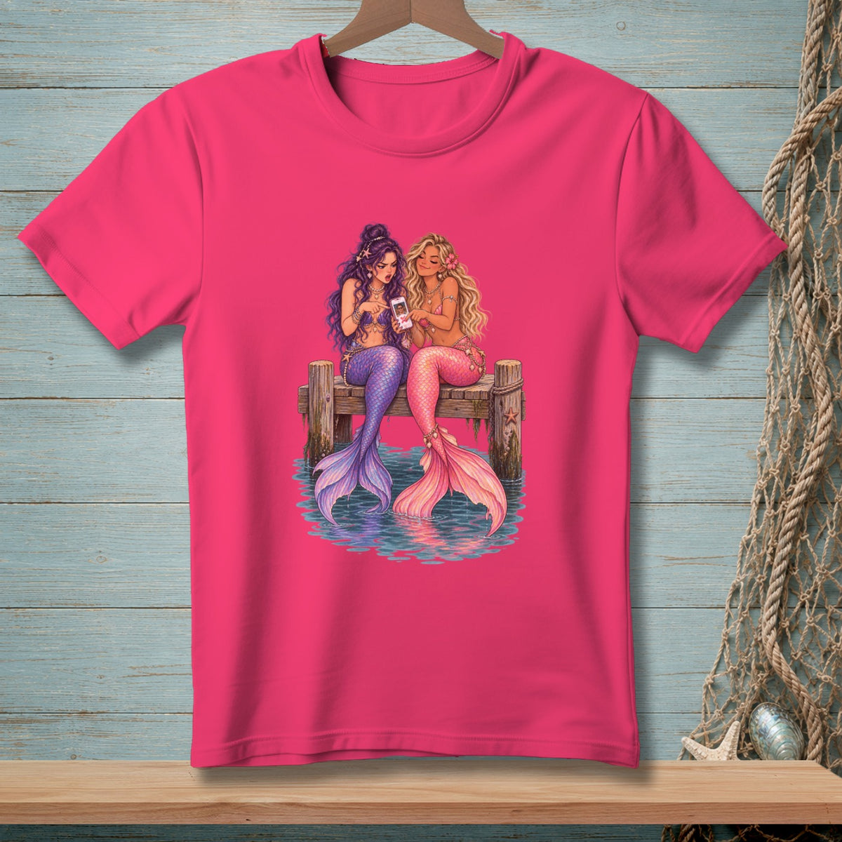 Two mermaids with purple and pink tails look at a smartphone on a wooden pier.