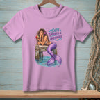 Pink t-shirt with a mermaid drinking from a coconut on a dock. Text says "Salty, Tangled & Unbothered."
