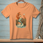 Coral t-shirt with a mermaid looking in a mirror held by a crab on a dock.