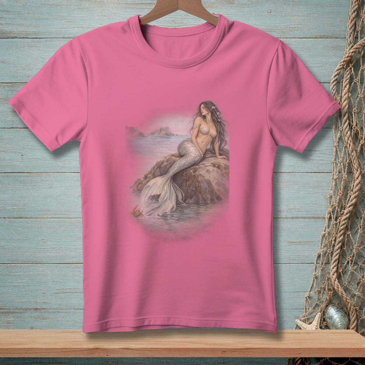 Pink t-shirt with an artistic print of a mermaid sitting on a rock by the sea.