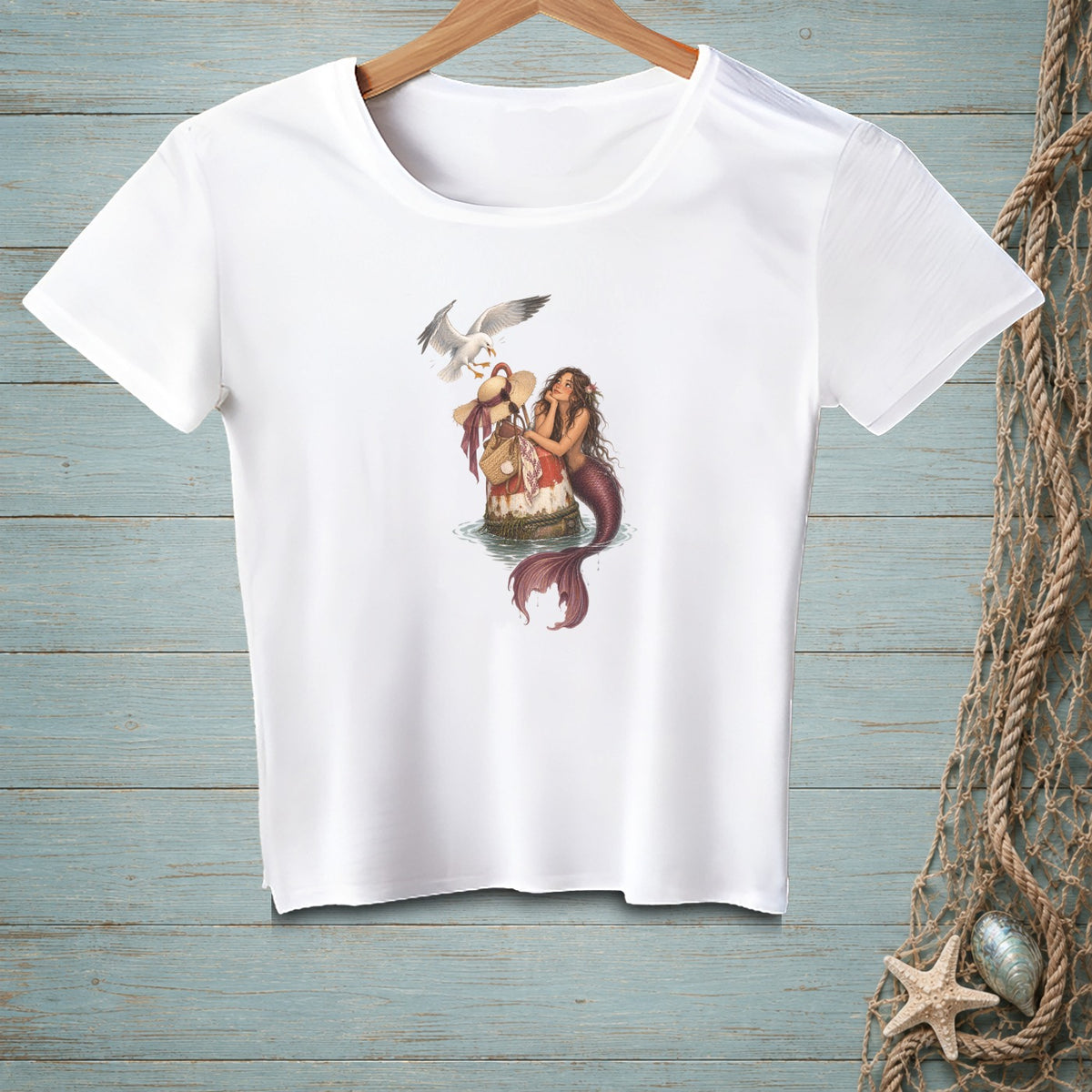 A white t-shirt hanging on a wooden wall features a graphic of a mermaid and seagull.