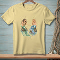 Light yellow t-shirt with two mermaids sitting on rocks in the ocean.