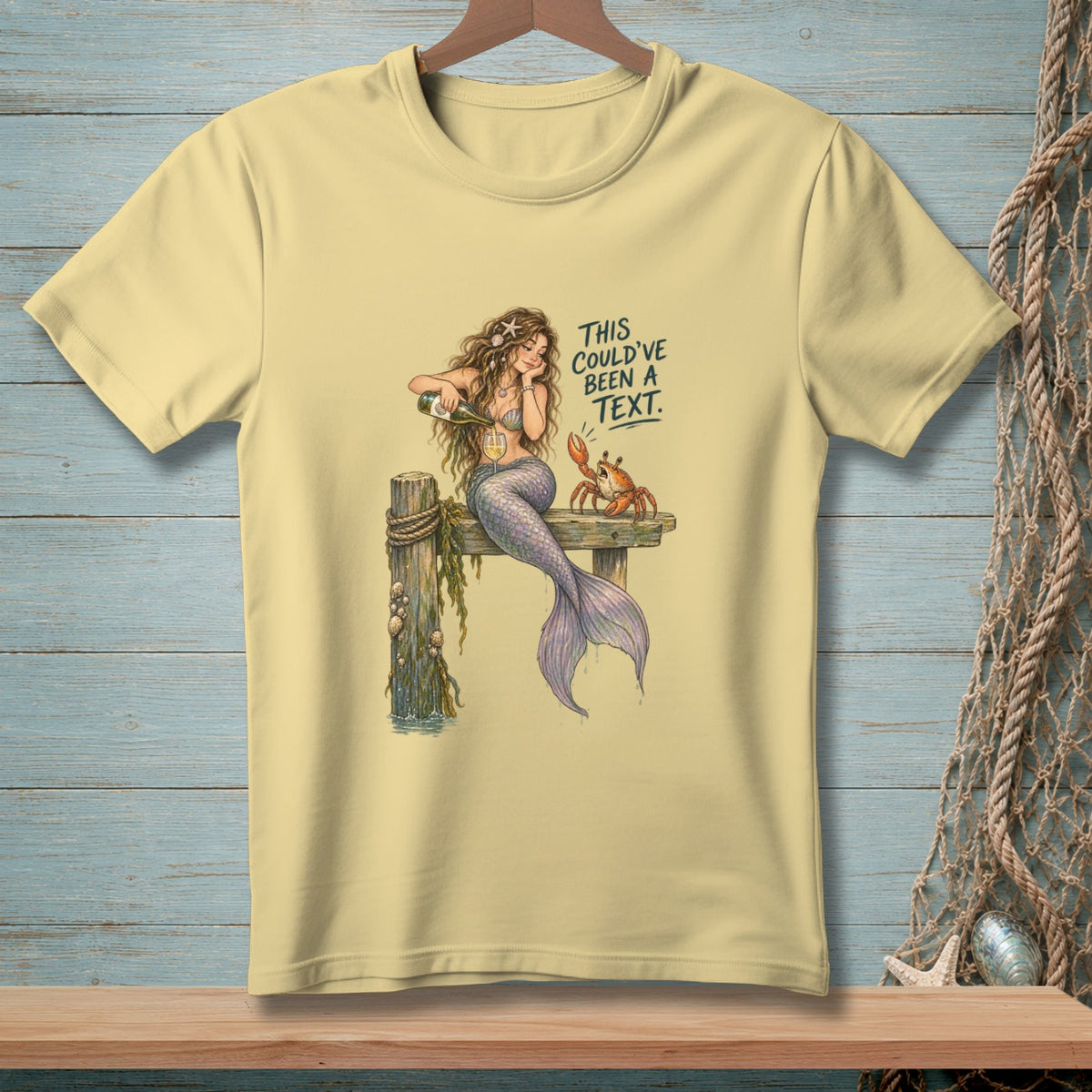 Light yellow t-shirt with a mermaid pouring champagne next to a crab, with text.