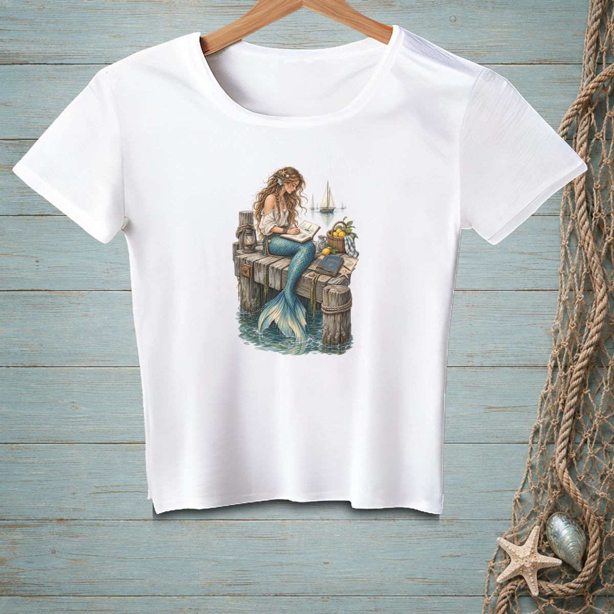 White t-shirt with a mermaid reading on a dock with sailboats in the background.