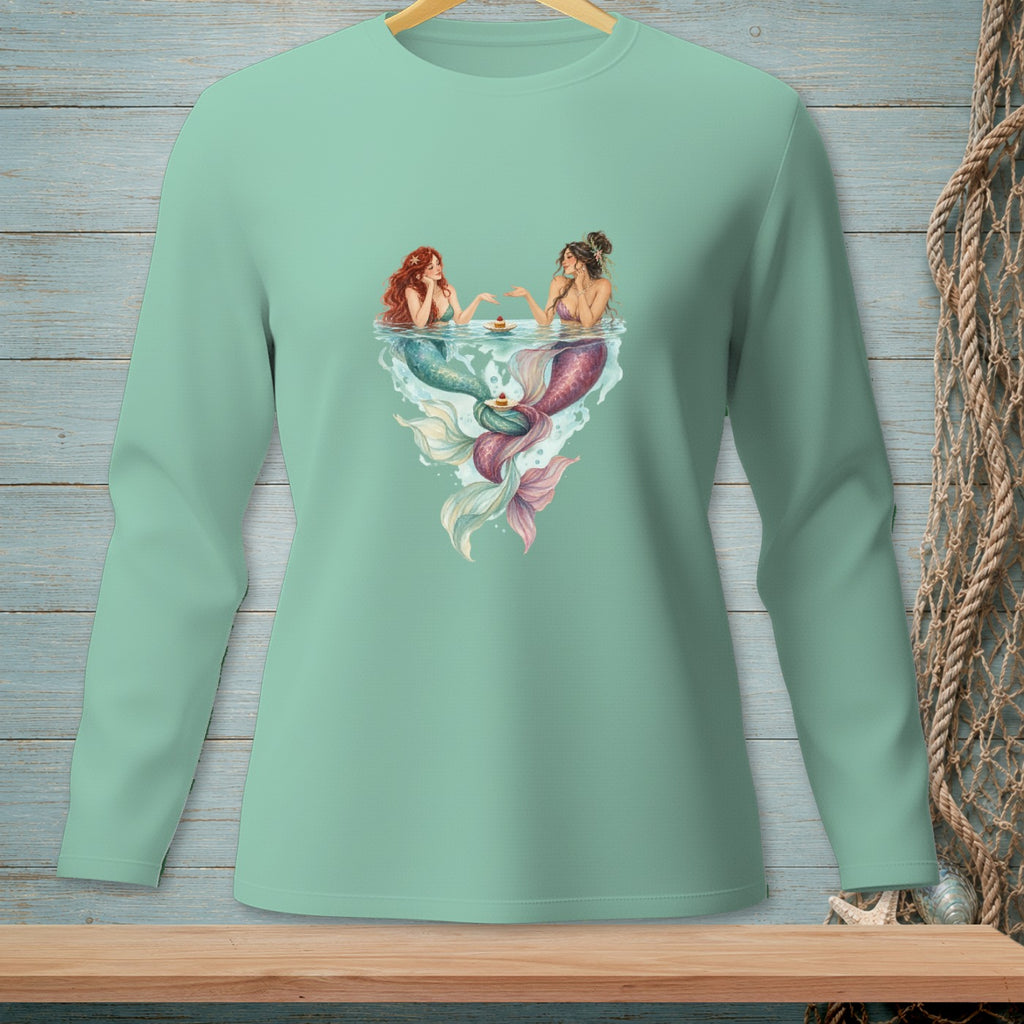 Two mermaids with colorful tails sit at a table of water with cakes.