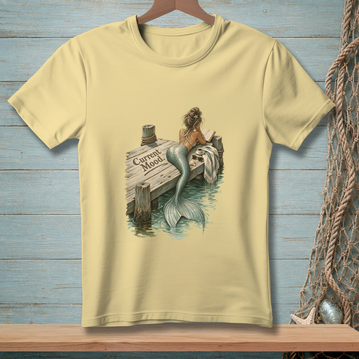 Yellow t-shirt with a mermaid reading on a dock, with text "Current Mood."