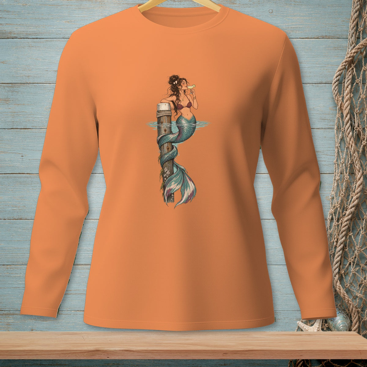 Orange long-sleeve shirt with a graphic of a mermaid on a wooden post drinking a cocktail.