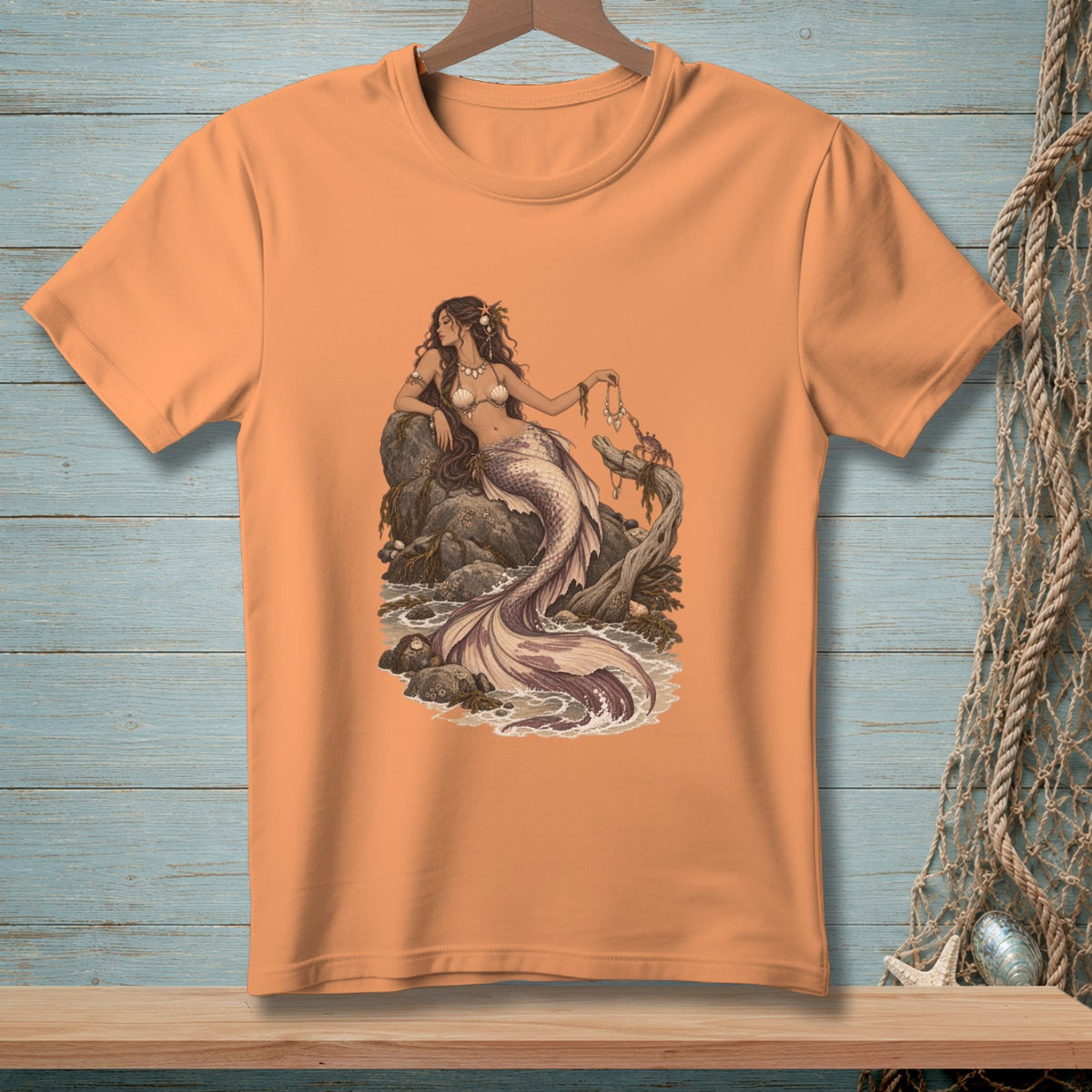 Peach t-shirt with an illustration of a mermaid sitting on rocks by the sea.