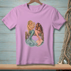 Two mermaids with colorful tails sit on a wooden dock, one looking in a mirror.