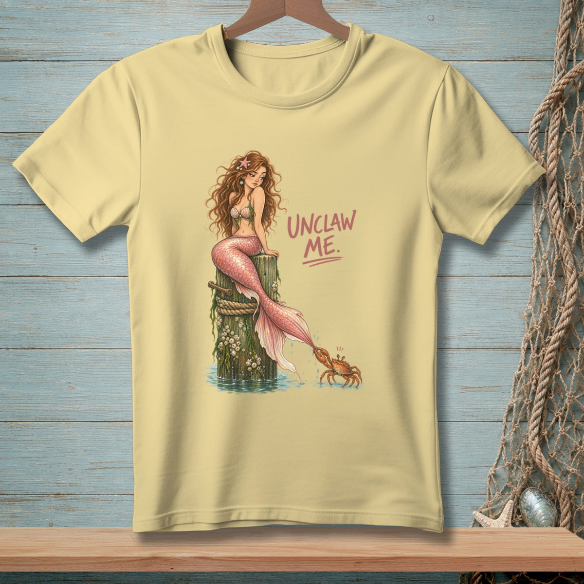Light yellow t-shirt with mermaid and crab illustration and "UNCLAW ME." text.