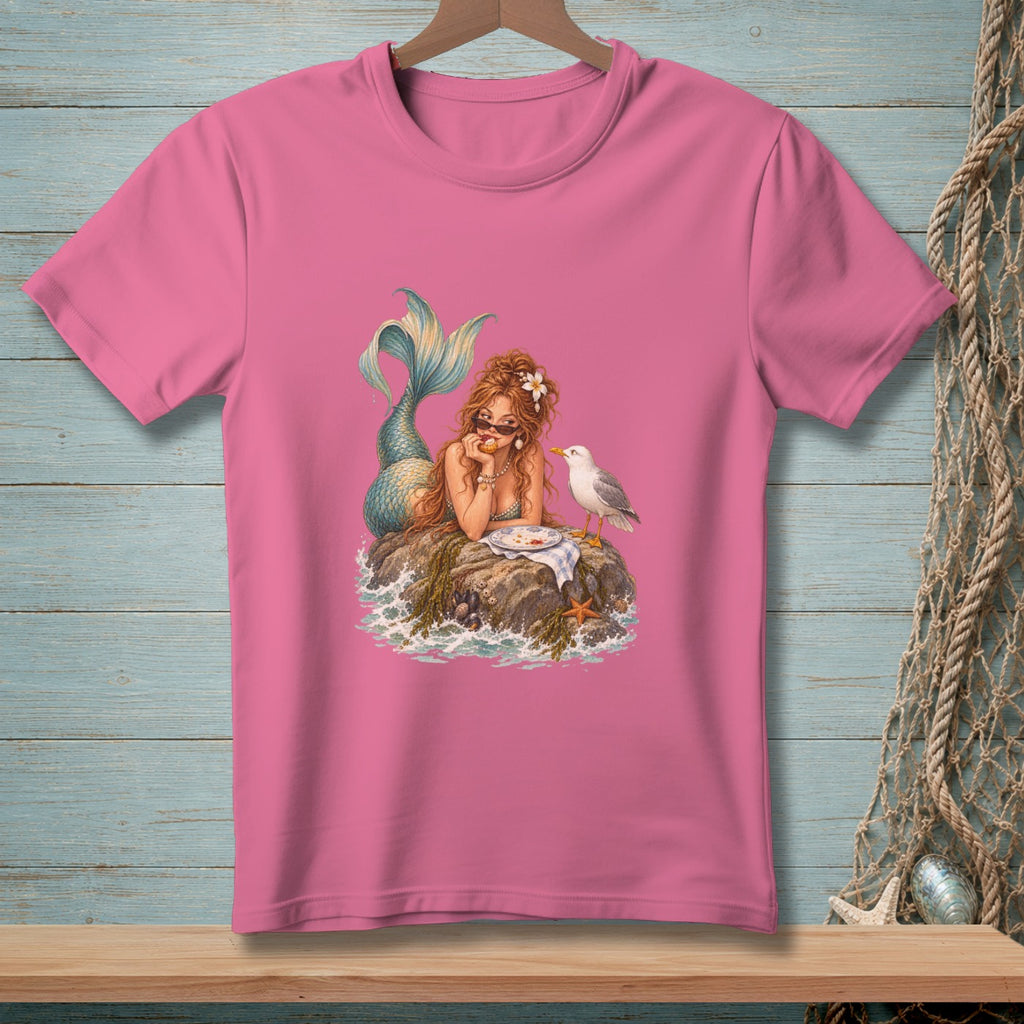 Pink t-shirt with a mermaid lounging on a rock with a seagull and a plate of food.