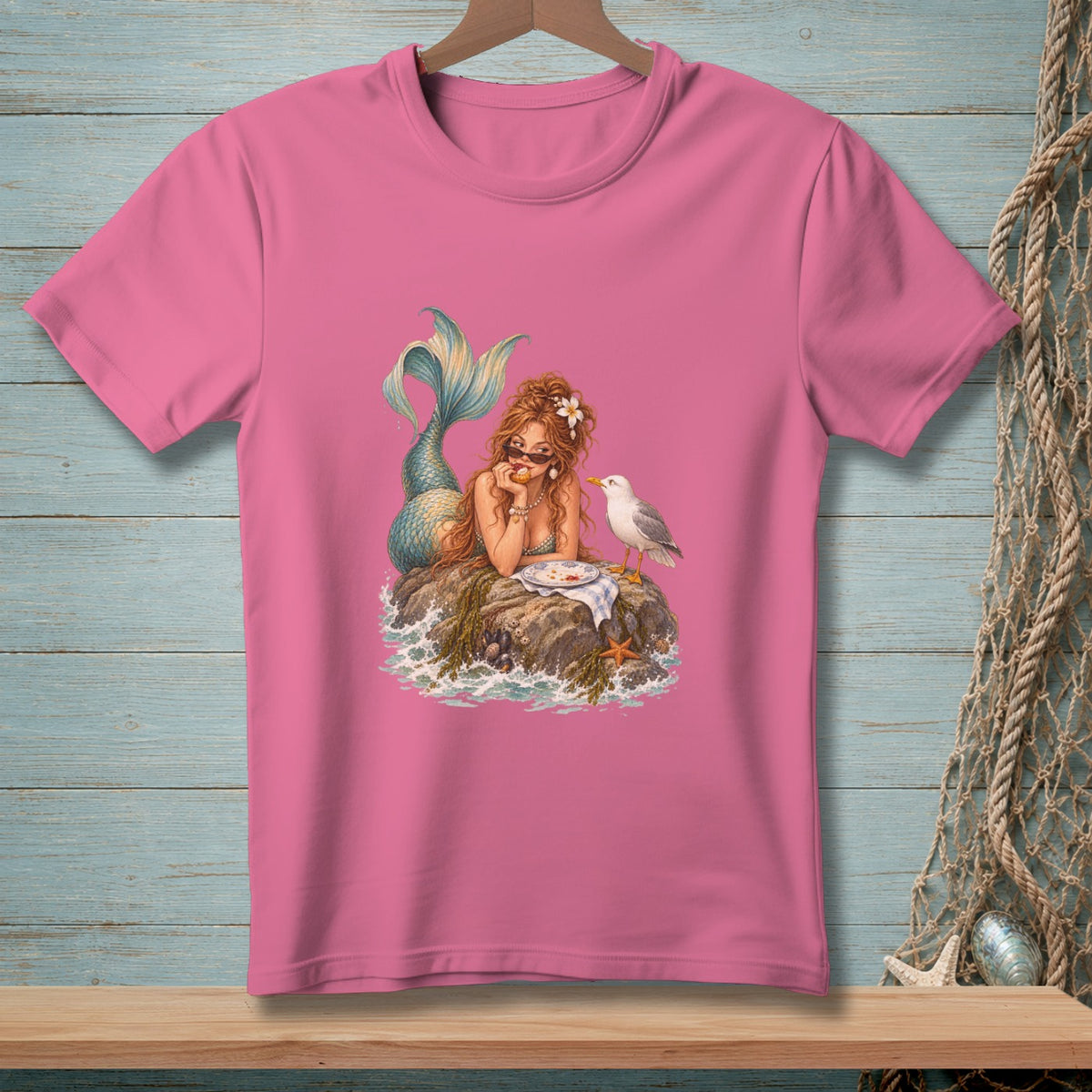 Pink t-shirt with a mermaid lounging on a rock with a seagull and a plate of food.