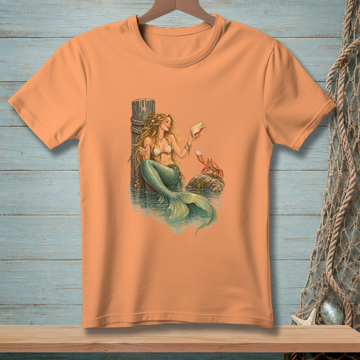 Peach t-shirt with a print of a mermaid holding a seashell next to a crab.